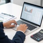 A business person analyzing financial charts and real estate data on a laptop, with calculator and documents spread out on a modern desk, professional office setting, focused atmosphere
