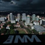 A dramatic aerial view of Kigali, Rwanda's capital, with modern buildings juxtaposed against a darkening sky. In the foreground, an IMF logo appears as a shadow. The image conveys economic uncertainty and crisis atmosphere.