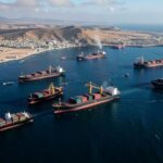A dramatic aerial view of oil tankers and cargo ships stalled at the Strait of Hormuz, with Middle Eastern coastline in the background, showing geopolitical tension blocking global trade routes.