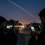 A dramatic scene in Tehran at dusk showing a civilian discreetly filming a military convoy with a smartphone, with missile trails visible in the distant night sky, reflecting the tension of citizen intelligence warfare.