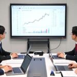 A modern Korean government office with policy documents showing stock market growth charts and KOSDAQ logos, officials discussing economic strategies around a table with laptops and financial reports.