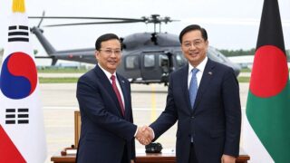 A symbolic handshake between South Korean and Bangladeshi officials with LH Mi-ru helicopters in the background, representing defense export diplomacy in 2026, professional business meeting atmosphere
