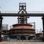 A symbolic image of an old, large industrial steel plant with a gate closing, representing the end of an era, with a modern city skyline in the background, conveying industrial change and history.