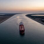 Aerial view of the Strait of Hormuz with oil tankers passing through, showing the narrow waterway between Iran and Oman, with flags of both countries visible on the shores, at sunset.