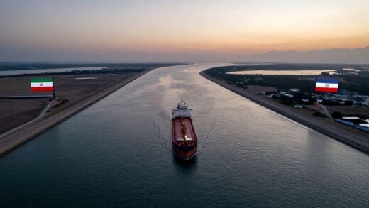 Aerial view of the Strait of Hormuz with oil tankers passing through, showing the narrow waterway between Iran and Oman, with flags of both countries visible on the shores, at sunset.